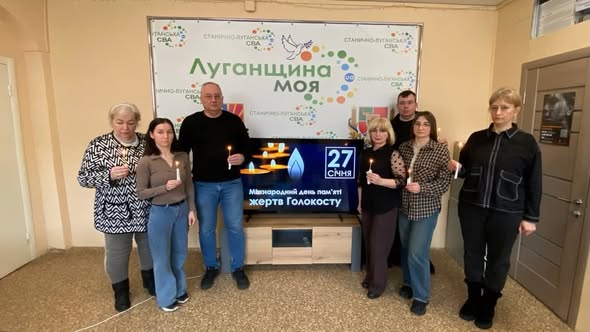 На зображенні показано групу з шести людей (три жінки та троє чоловіків), які стоять у приміщенні та тримають у руках запалені свічки. Вони стоять перед великим банером із написом «Луганщина моя». Під банером розташований телевізор, на екрані якого відображається напис «Міжнародний день пам'яті жертв Голокосту» та дата «27 СІЧНЯ» разом із зображенням свічок