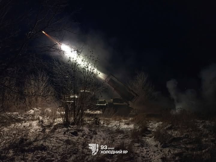 Зображення показує нічну сцену з військовою технікою (пусковою установкою), яка веде вогонь. На темному тлі видно яскравий спалах пострілу та димовий слід від ракети, що летить угору вліво. Земля вкрита снігом та сухою рослинністю