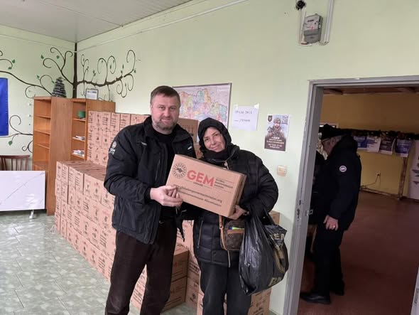 Зображення є фотографією двох людей, ймовірно, волонтерів, які тримають картонну коробку з гуманітарною допомогою. На коробці видно логотип «GEM» (Global Empowerment Mission) та напис "UKRAINE", а також адресу веб-сайту globalempowermentmission.org