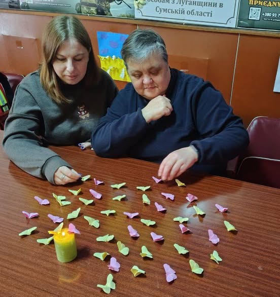 На зображенні дві жінки сидять за дерев'яним столом і працюють із численними маленькими паперовими фігурками (схожими на метеликів або сердечка) рожевого та світло-зеленого кольорів. Фігурки розкидані по поверхні столу. На передньому плані ліворуч стоїть невелика запалена свічка у зеленій підставці.
