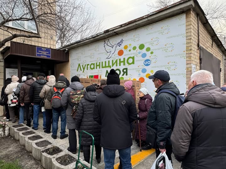 На зображенні видно чергу людей, одягнених у зимовий одяг, які стоять перед будівлею з великим банером із написом «Луганщина моя», ймовірно, очікуючи на отримання гуманітарної допомоги або послуг.