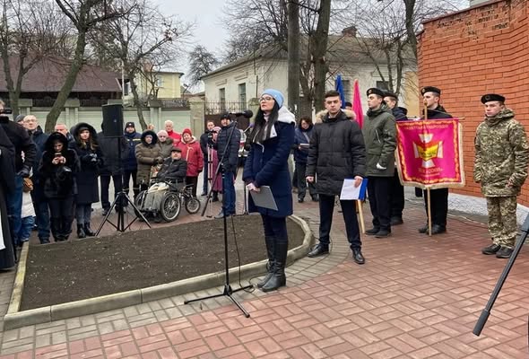 На зображенні видно натовп людей, які зібралися на офіційній церемонії на відкритому повітрі, де жінка виступає біля мікрофона, а молоді люди у формі тримають прапори.