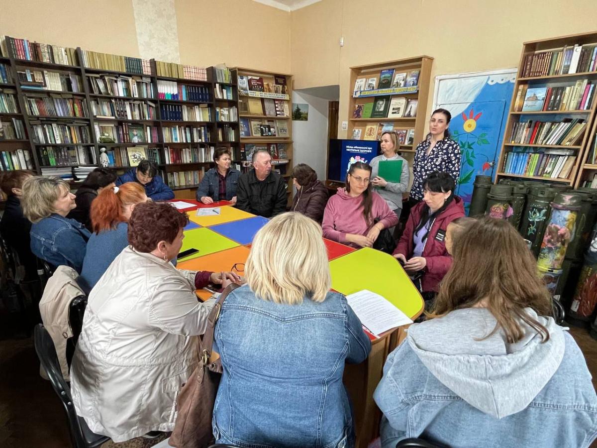Група з приблизно десяти людей, переважно жінок, сидить навколо яскравого столу в бібліотеці, де на задньому плані видно книжкові полиці та предмети, схожі на військову техніку, слухаючи жінку, яка стоїть біля дошки