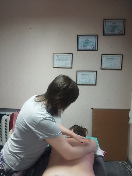 Зображення показує, як фахівець проводить сеанс масажу спини клієнту на масажному столі, при цьому на стіні позаду висять декілька сертифікатів або дипломів у рамках