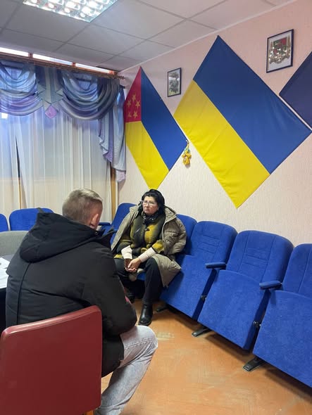Зображення показує двох людей, що сидять обличчям один до одного під час зустрічі в офіційній кімнаті, прикрашеній прапорами України та регіональним прапором, а також рядами синіх крісел.