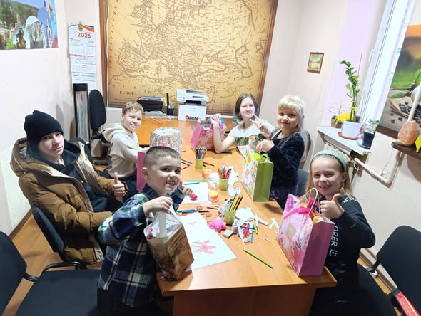 Шестеро дітей різних вікових груп радісно сидять за столом в офісі, де на стіні висить велика карта, тримають подарункові пакети та демонструють великі пальці вгору.