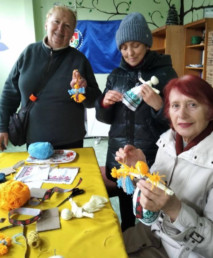 На фото зображено трьох жінок, які під час волонтерського майстер-класу в Закарпатті власноруч створюють традиційні українські ляльки-мотанки та патріотичні обереги з жовто-блакитної пряжі та вишитого полотна, символізуючи єдність та незламність національного духу