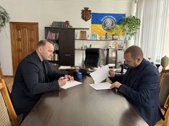 Двоє чоловіків підписують документи за столом в офісі, де на стіні висить український прапор.