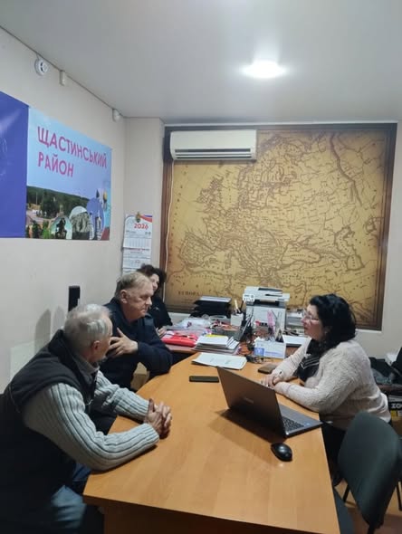 Двоє чоловіків та жінка сидять за столом в офісі, де на стіні висить карта та стоїть ноутбук.