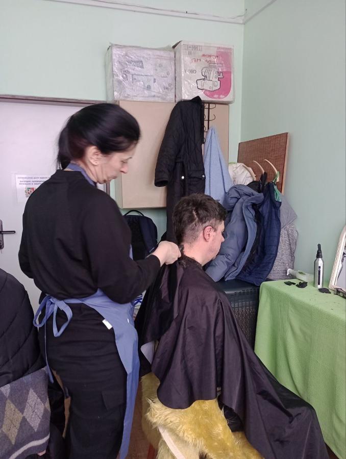 Жінка в чорній кофті та блакитному фартуху стриже чоловіка в чорному перукарському пеньюарі в невеликому приміщенні, де на задньому плані видно складені коробки, підвішений верхній одяг та перукарське приладдя