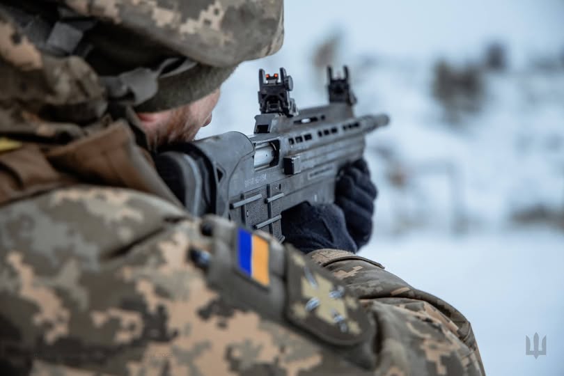 На світлині зображений український військовослужбовець у піксельному камуфляжі, який цілиться з автоматичної гвинтівки «Малюк» (Вулкан-М) на тлі засніженого краєвиду. На плечі бійця чітко видно шеврон із національним прапором та тактичним знаком, що підкреслює його приналежність до Сил оборони України