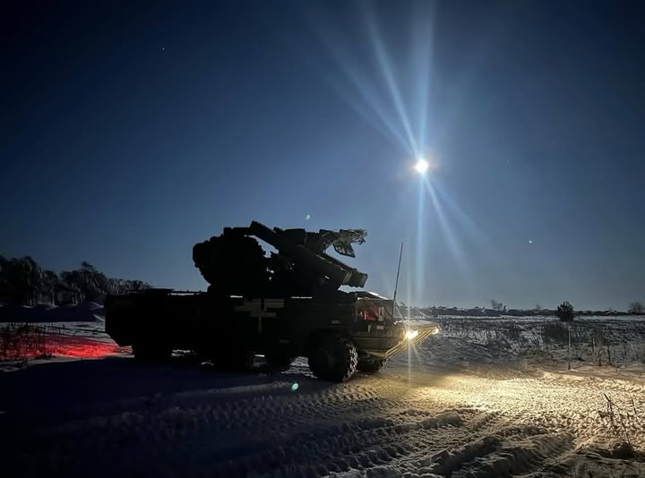 Військова вантажівка, оснащена зенітно-ракетним комплексом, стоїть у засніженому полі в нічний час, освітлена яскравим місяцем та фарами