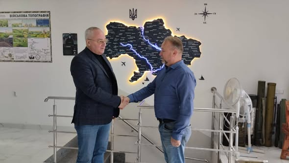 На зображенні представлено двоє чоловіків у приміщенні, які тиснуть один одному руки на фоні стіни з картою України та плакатом з військової топографії. Чоловік ліворуч одягнений у темно-синій піджак, чорний гольф та джинси, а чоловік праворуч — у синю сорочку та джинси. Зустріч відбувається біля сходової клітки з металевими поручнями, а поруч на підлозі стоять предмети, схожі на тубуси для зброї або обладнання