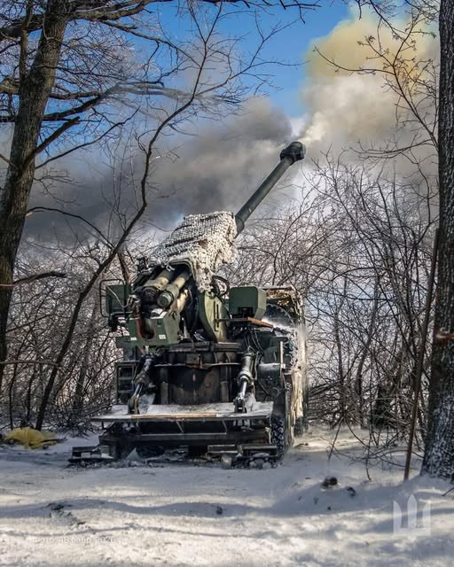 Українська самохідна артилерійська установка 2С22 «Богдана» веде вогонь по позиціях противника в зимовому лісі. Ствол гармати піднятий вгору, з нього виривається дим після пострілу; установка частково вкрита білою маскувальною сіткою.