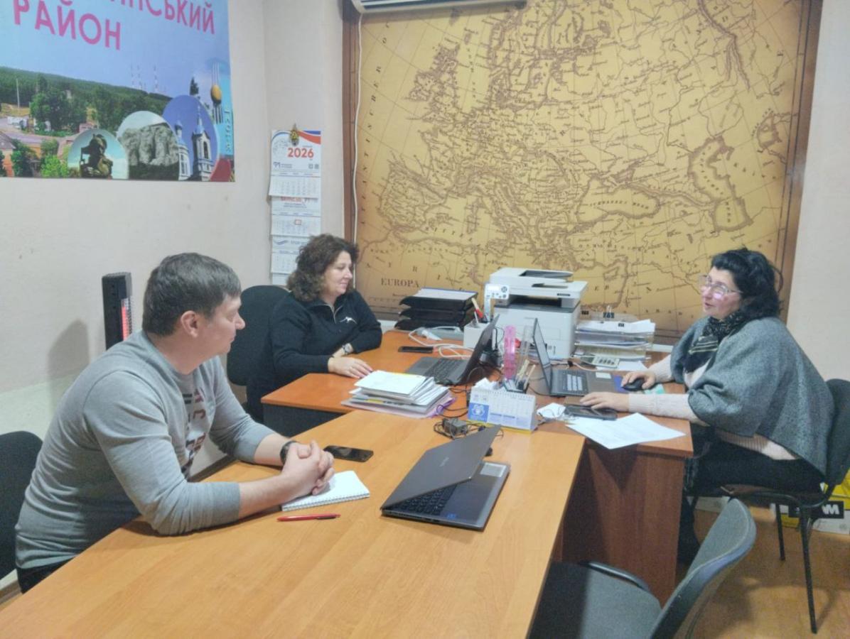 Три людини, двоє чоловіків та жінка, сидять за дерев'яним столом в офісі, де на стіні висить велика карта Європи та плакат з написами