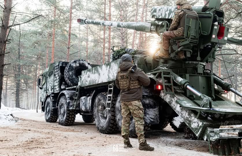 На цьому зображенні українські військовослужбовці працюють із вітчизняною самохідною артилерійською установкою (САУ) 2С22 «Богдана» у зимовому лісі. Один боєць розрахунку перебуває на сидінні навідника біля гармати, а інший стоїть поруч із великогабаритною колісною платформою, ймовірно, на шасі Tatra