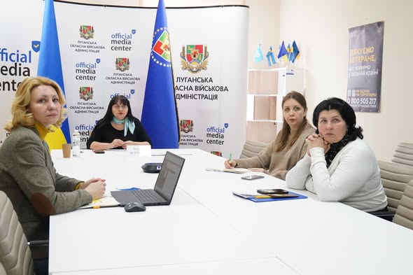 Чотири жінки проводять офіційну зустріч або брифінг у приміщенні «Official Media Center» Луганської обласної військової адміністрації. Вони сидять за великим білим столом, на якому стоять ноутбук та документи. На задньому плані розміщено прапор Луганської області, логотипи медіацентру та інформаційний плакат для ветеранів
