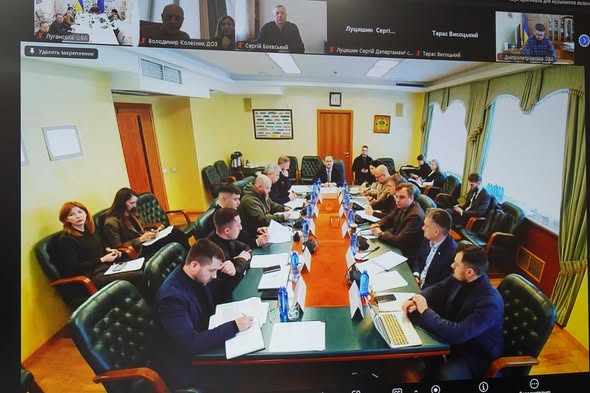 На екрані ноутбука відображено робочу зустріч: група посадовців сидить за великим столом у залі засідань, а у верхній частині вікна видно іконки учасників відеоконференції. На столі лежать папери, ноутбуки та пляшки з водою