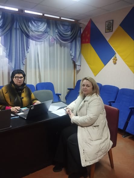 Дві жінки проводять робочу зустріч в офісному приміщенні. Одна сидить за столом із ноутбуком, інша — поруч із документами. На стіні позаду розміщені прапори України та Луганської області
