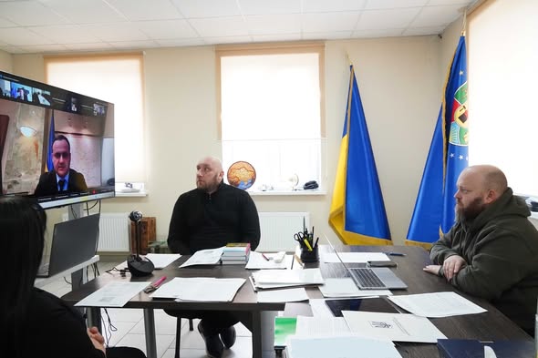За столом сидить двоє чоловіків у темному одязі та худі кольору хакі, а також жінка (вид зі спини). На великому настінному екрані відображається відеодзвінок з іншим учасником