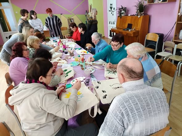 Група людей різного віку, переважно похилого, зібралася в яскравій кімнаті для творчого майстер-класу. Вони сидять за довгим столом і розмальовують фарбами екосумки. На фоні видно піаніно, картини на стінах та інших учасників, які спостерігають за процесом.