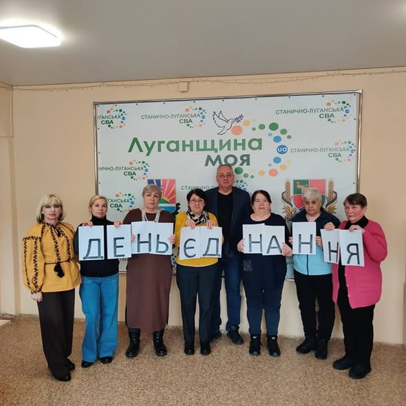 Група людей стоїть у приміщенні на фоні банера з написом «Луганщина моя» та логотипами Станично-Луганської СВА. У руках вони тримають аркуші паперу, які разом утворюють напис «ДЕНЬ ЄДНАННЯ».
