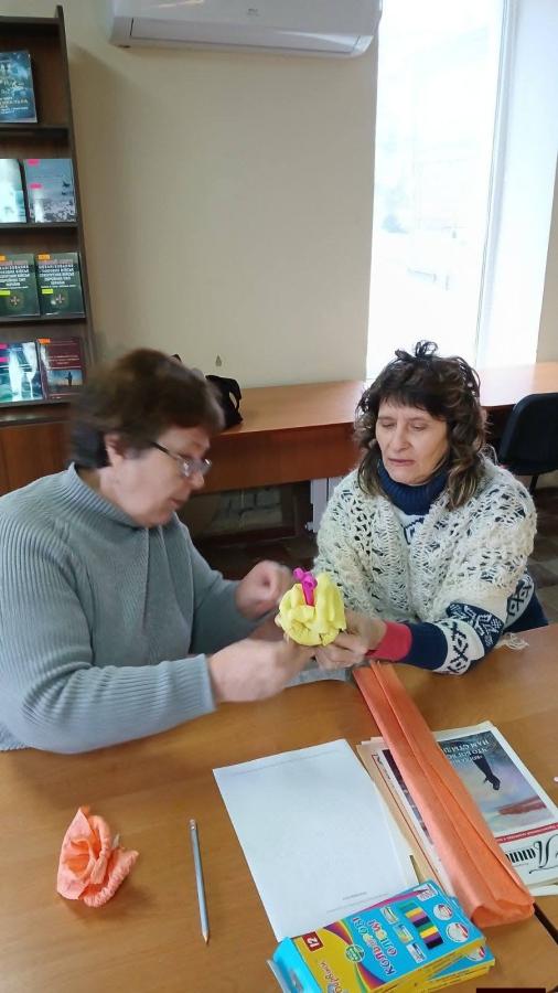 Дві жінки старшого віку, які зосереджено працюють над створенням виробу. Одна з них показує іншій, як правильно формувати деталь