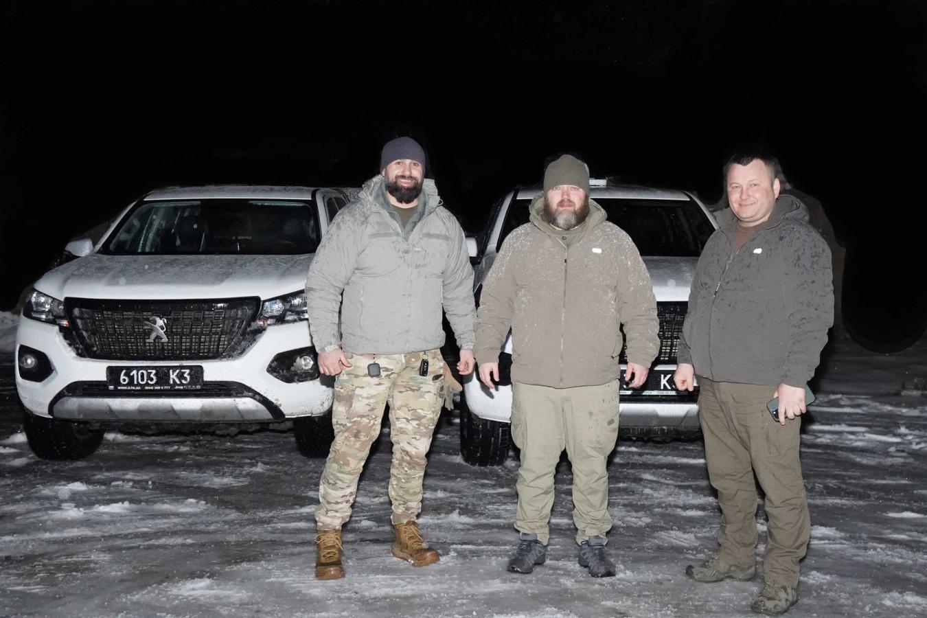 На зображенні — троє чоловіків у тактичному та повсякденному одязі, які стоять на снігу на фоні двох білих пікапів