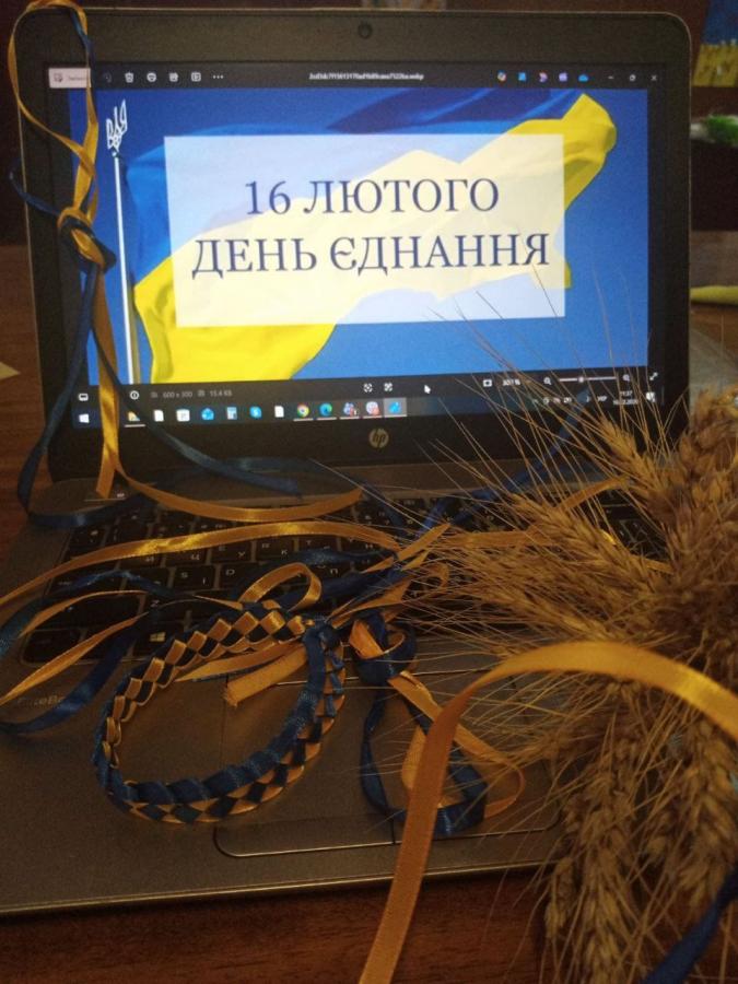 На ноутбуці відкрите зображення з написом «16 ЛЮТОГО ДЕНЬ ЄДНАННЯ» на фоні українського прапора. Перед клавіатурою лежать синьо-жовті стрічки, плетений браслет і пучок колосків пшениці.