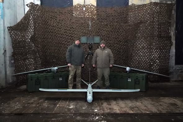 На фото зображено двох чоловіків, які стоять поруч із безпілотними авіаційними комплексами (БпАК)