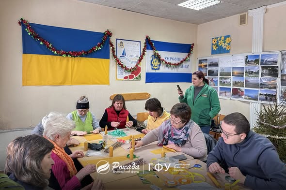 Група людей на творчому майстер-класі від Благодійного фонду «Рокада». Учасники сидять за столом у світлій кімнаті, прикрашеній українськими прапорами, та виготовляють вироби ручної роботи