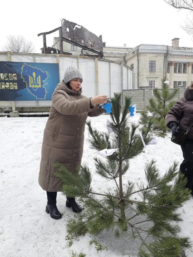 Жінка в подовженому коричневому пуховику та сірій в'язаній шапці розвішує на невелику сосну синіх та білих паперових ангелів. Ці фігурки є символом акції «Ангели пам'яті», яка традиційно проводиться в Україні до Дня Героїв Небесної Сотні (20 лютого)