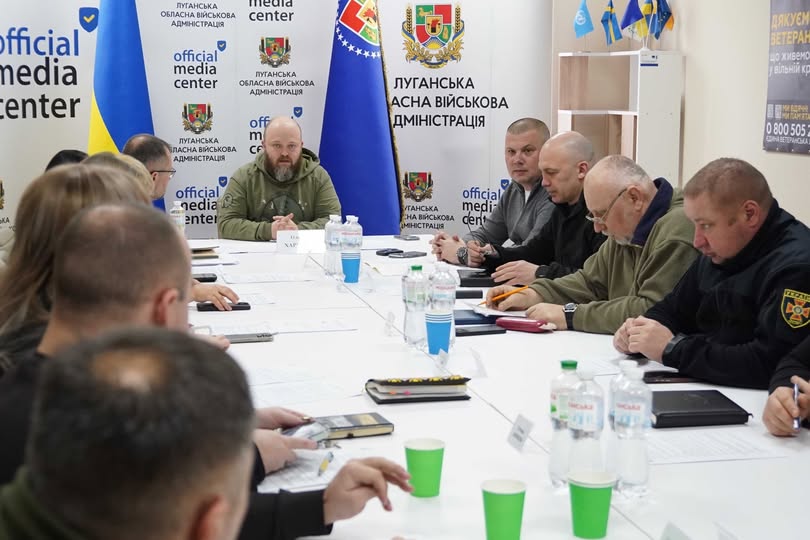 За довгим білим столом зібралася група людей. У центрі (на фоні прапорів) — чоловік у військовій формі оливкового кольору, який, ймовірно, головує на зустрічі. Праворуч від нього сидять представники різних відомств, зокрема рятувальник ДСНС (у чорній формі з нашивкою)