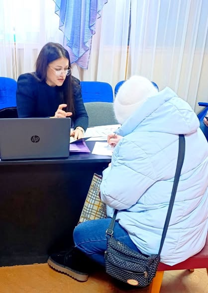 За столом сидить жінка-спеціаліст у діловому одязі та окулярах, вона щось пояснює та вказує на документи. Навпроти неї, спиною до камери, сидить відвідувачка у світлому зимовому одязі (пуховику та шапці), яка в цей момент підписує папери