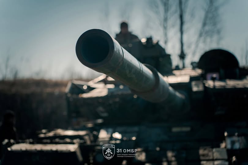 На фото зображено крупний план ствола гармати українського танка 31-ї окремої механізованої бригади, спрямованого вперед на тлі розмитого пейзажу та екіпажу