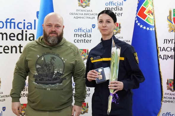 Чоловік у худі кольору хакі та жінка у формі ДСНС із квітами й нагородою позують на фоні банера Луганської обласної військової адміністрації