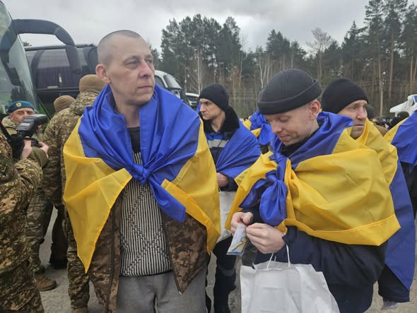 На фото зображено звільнених з полону українських захисників, які стоять на вулиці, загорнуті в національні прапори України