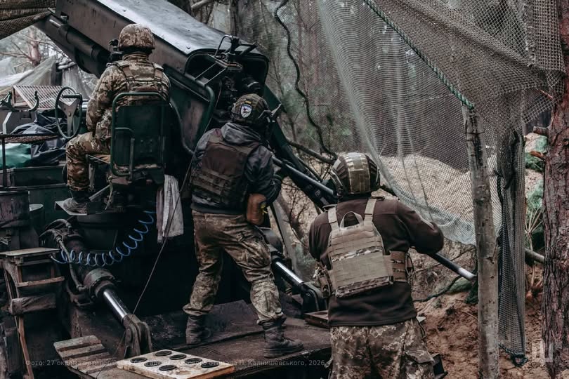 Троє українських артилеристів у повному спорядженні працюють біля розгорнутої артилерійської установки під маскувальною сіткою в лісистій місцевості.