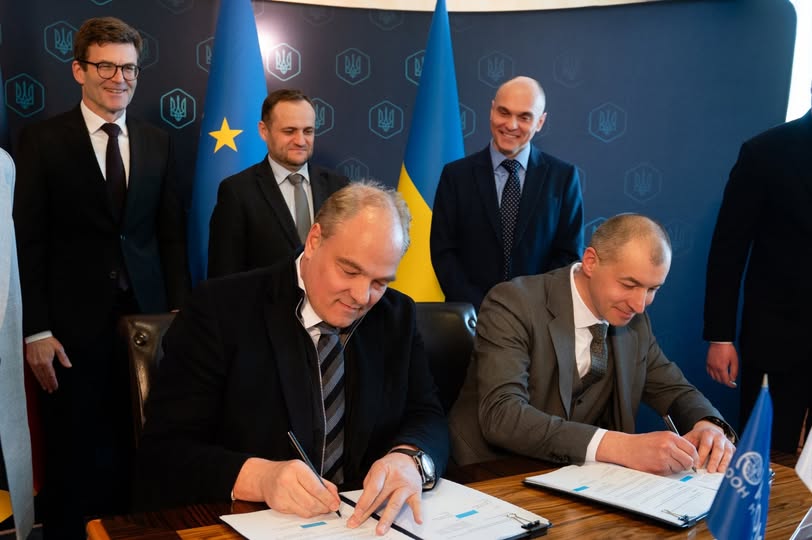 На офіційному заході двоє чоловіків у ділових костюмах підписують документи за столом, у той час як за ними стоять інші офіційні особи на фоні прапорів України та Європейського Союзу