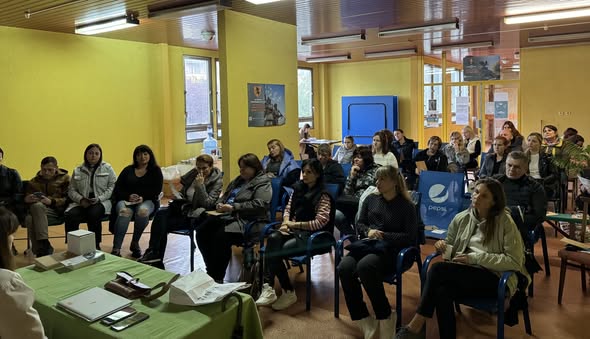Група людей уважно слухає доповідача під час інформаційної зустрічі або тренінгу в жовтій аудиторії, де на столі перед ними розкладені блокноти та матеріали для записів.