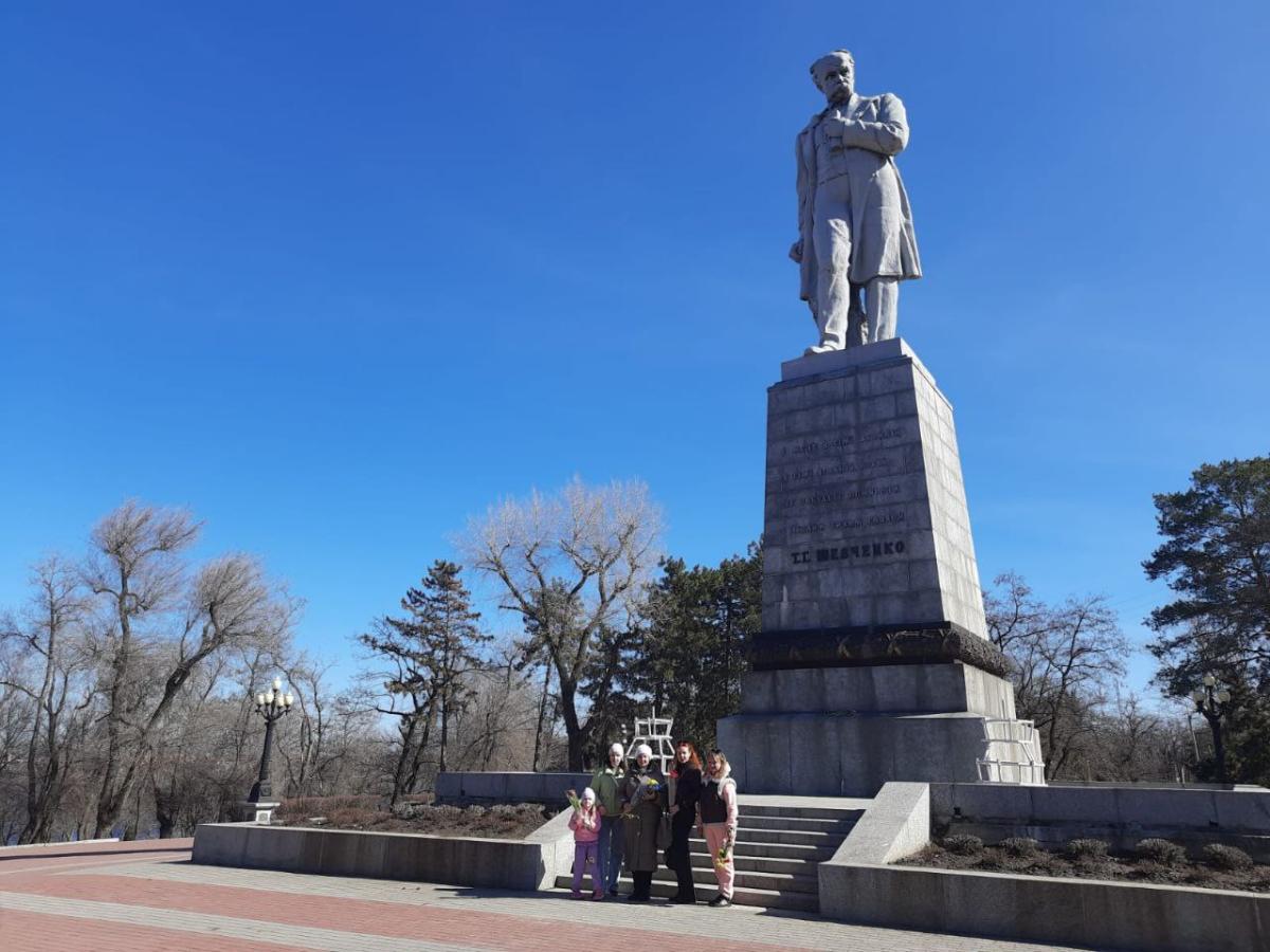 Група людей позирує для спільного фото біля підніжжя величного пам'ятника Тарасу Шевченку на Монастирському острові в Дніпрі за ясної сонячної погоди.