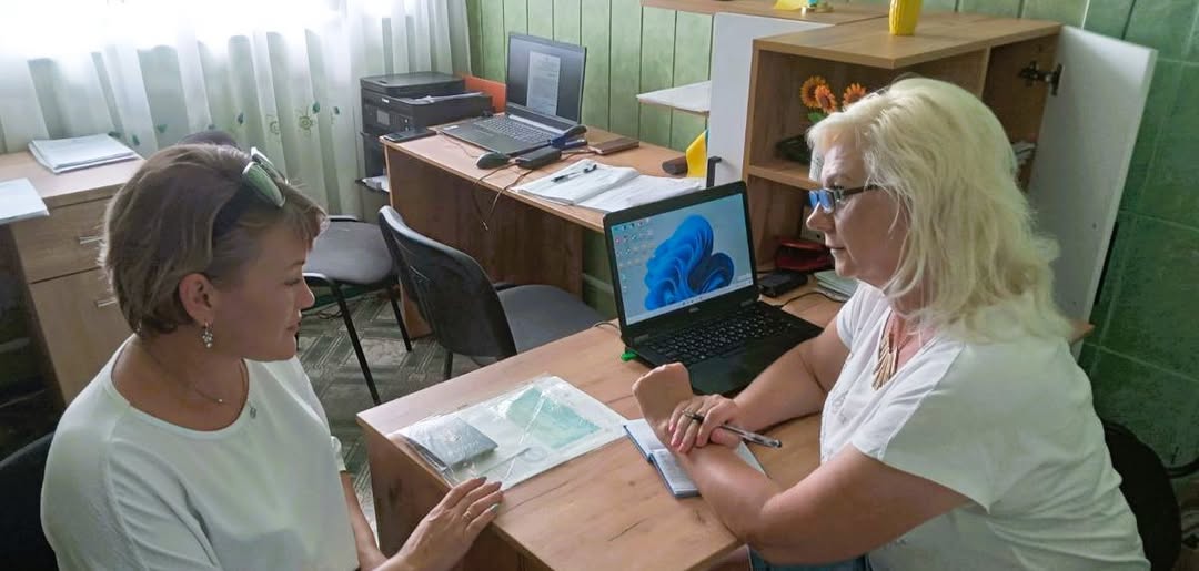Дві жінки в білих футболках сидять за робочими столами одна навпроти одної в офісному приміщенні зі світло-зеленими стінами та ноутбуками, обговорюючи документи під час робочого процесу.