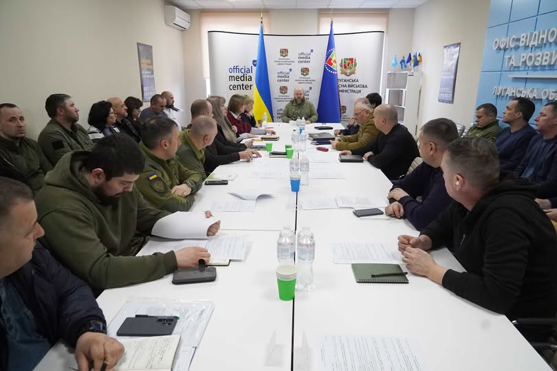На світлині зображено офіційне засідання групи посадовців та військових, які сидять за довгим білим столом у конференц-залі «Official Media Center» на фоні державних прапорів України