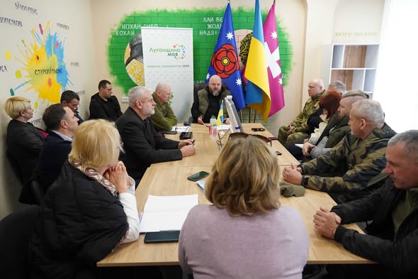 На світлині зафіксовано робочу зустріч групи людей за довгим дерев’яним столом у світлому офісному приміщенні, прикрашеному державною символікою та банером «Луганщина моя». Учасники, серед яких військові та цивільні, зосереджено слухають доповідача в центрі, що свідчить про обговорення важливих соціальних чи адміністративних питань регіону