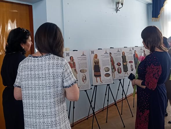 Троє жінок розглядають виставку інформаційних постерів на мольбертах, що демонструють різноманітні зразки традиційного українського вбрання з ілюстраціями та описом їхніх особливостей.