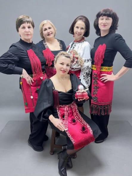 П'ять усміхнених жінок позують для групового фото в автентичному вбранні, що поєднує сучасний чорний одяг із традиційними українськими елементами — вишиванками та яскраво-червоними декоративними фартухами з вишивкою та бахромою