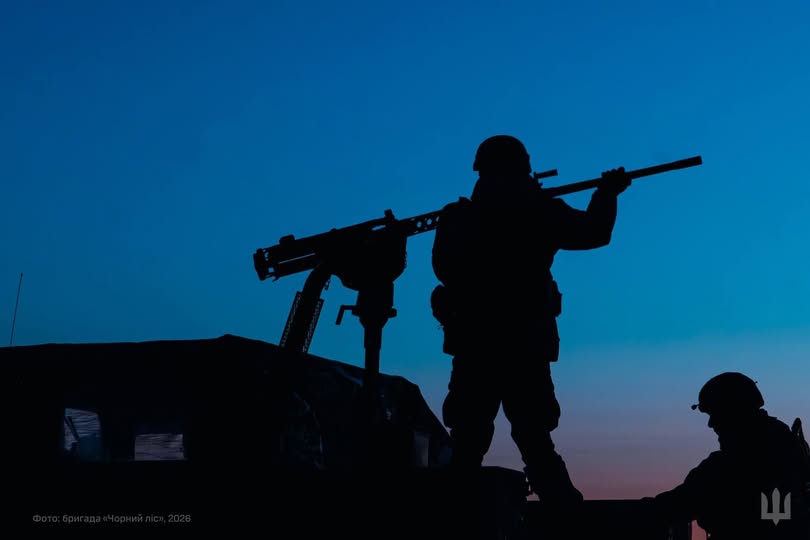 На світлині зображено темні силуети двох українських військових та великокаліберного кулемета на фоні глибокого синього неба в сутінках. Один із бійців стоїть біля турелі, перевіряючи готовність зброї, встановленої на військовому автомобілі бригади «Чорний ліс»