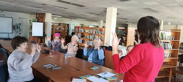 На світлині зображена група жінок, які сидять за великим столом у просторій бібліотеці та повторюють жести руками за жінкою в червоному светрі, що стоїть до камери спиною. Учасниці зустрічі, серед яких є люди старшого віку, зосереджено підняли руки вгору, а на фоні видно численні книжкові полиці, фліпчарт та навчальне приладдя на столі.