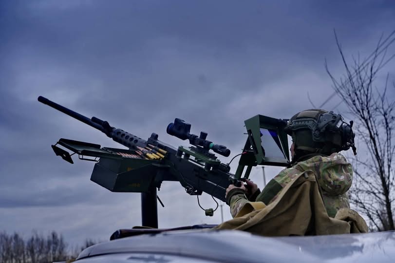 Військовослужбовець у повному бойовому спорядженні керує встановленим на пікапі великокаліберним кулеметом, оснащеним сучасним оптичним прицілом та виносним монітором для точного наведення на фоні похмурого неба.