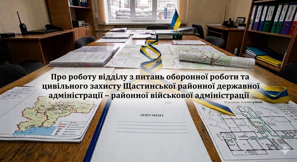 На великому дерев'яному столі в кабінеті Щастинської районної військової адміністрації розкладені топографічні карти, схеми евакуації та офіційні документи, обрамлені синьо-жовтими стрічками, тоді як на задньому плані через вікно видно захисні укріплення з мішків із піском